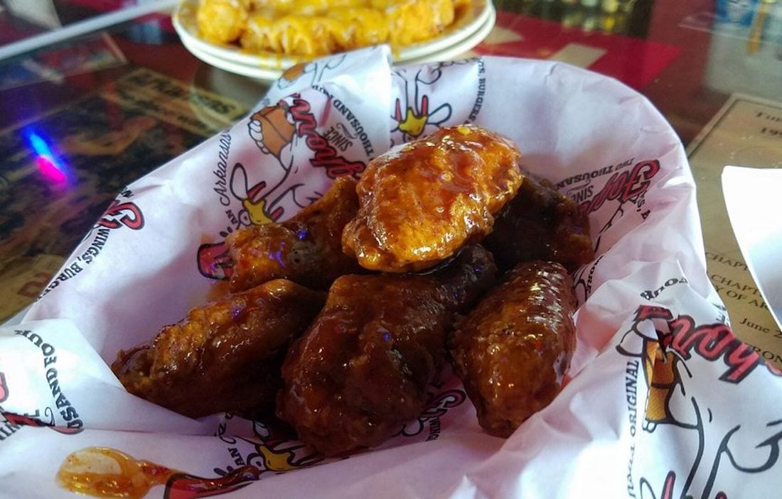 A basket lined with branded paper holds some of the best chicken wings, saucy and tempting. In the background, a blurry plate of fries sits on a reflective table next to a laminated menu.