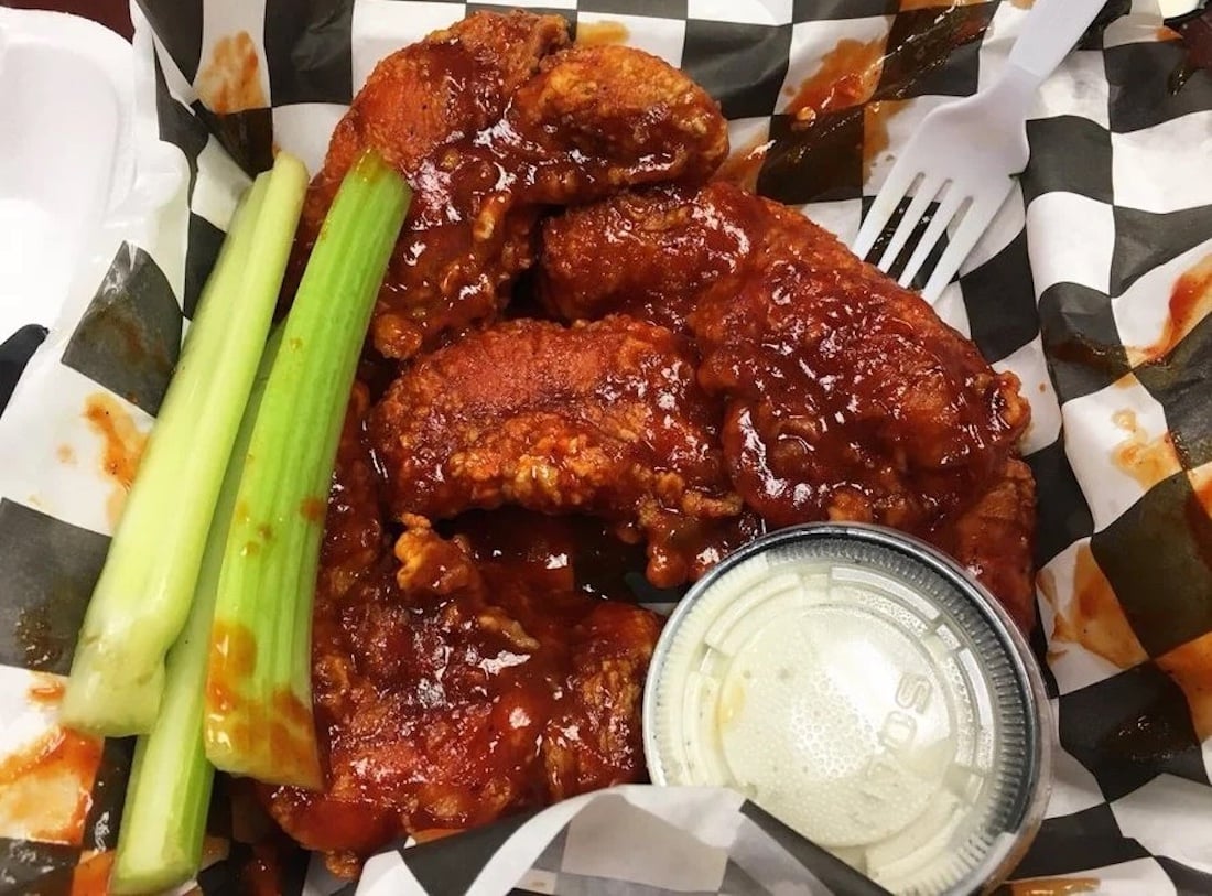 Chicken tenders coated in a spicy red sauce sit next to celery sticks and a cup of ranch dressing, served in a paper-lined basket with a plastic fork—perfect for fans of the best chicken wings.