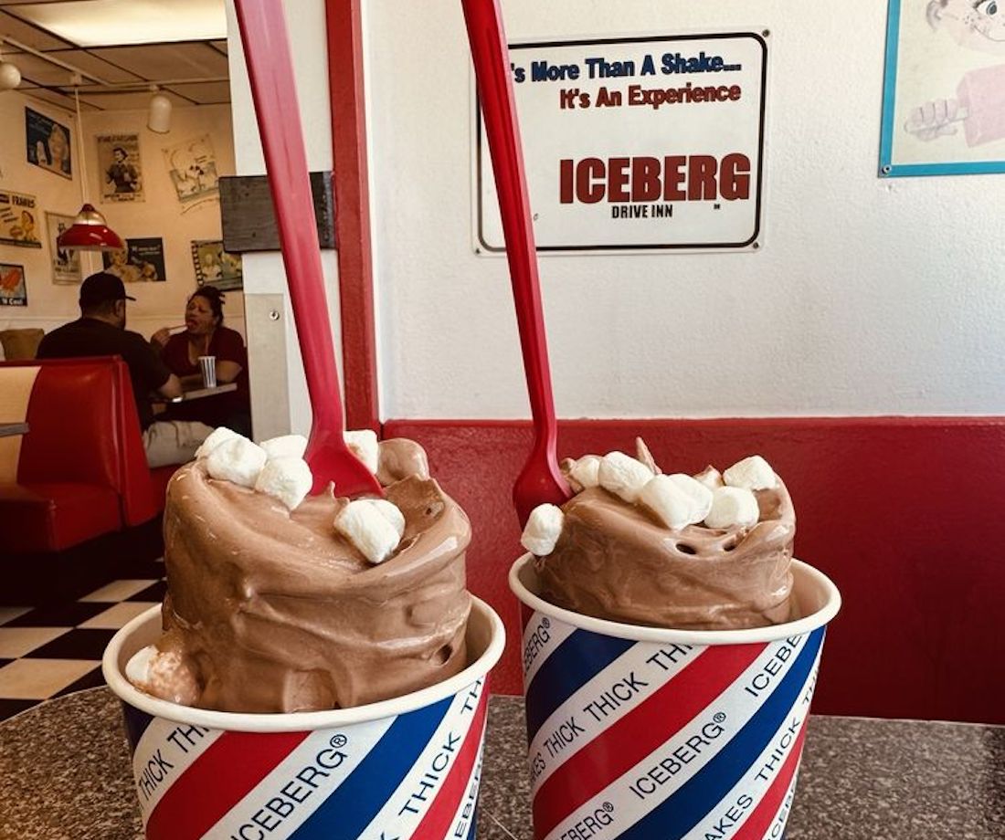Two large cups of thick chocolate milkshake topped with mini marshmallows and red spoons sit on a table in a retro-style diner with a “ICEBERG” sign on the wall and people seated in a booth in the background.