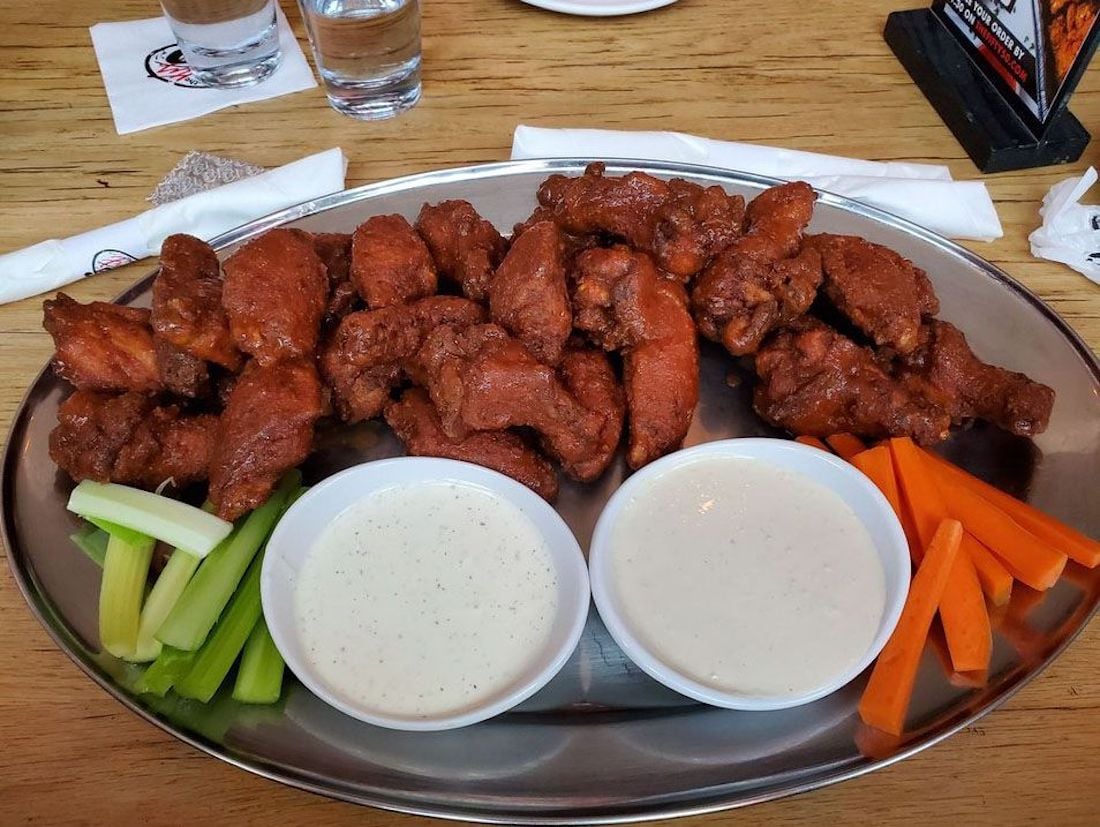A metal platter filled with the best chicken wings—buffalo-style—served with two bowls of ranch dipping sauce and sides of celery and carrot sticks, all on a wooden table.