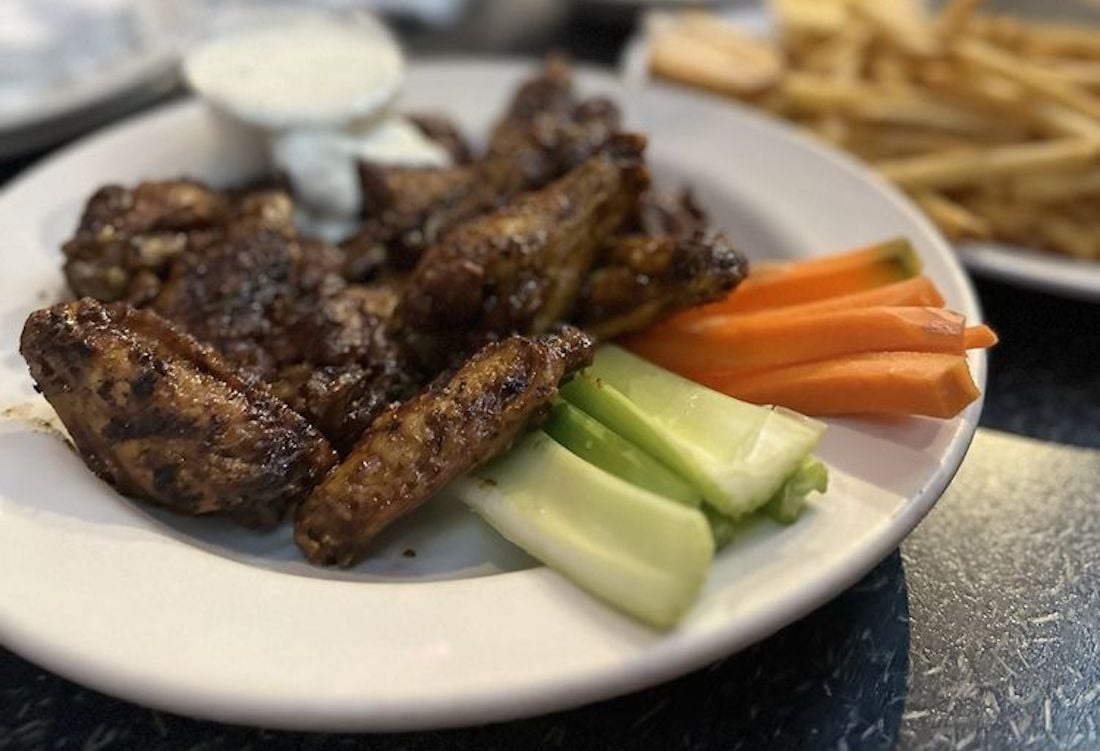 A plate of the best chicken wings, grilled to perfection, with a cup of ranch dip, served alongside crisp celery and carrot sticks. A plate of French fries is blurred in the background.
