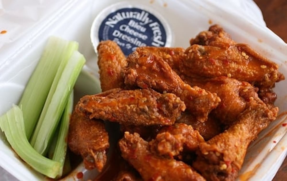 A takeout container holds the best chicken wings tossed in spicy buffalo sauce, accompanied by crisp celery sticks and a small cup of blue cheese dressing from Jamal's Buffalo Wings in Atlanta