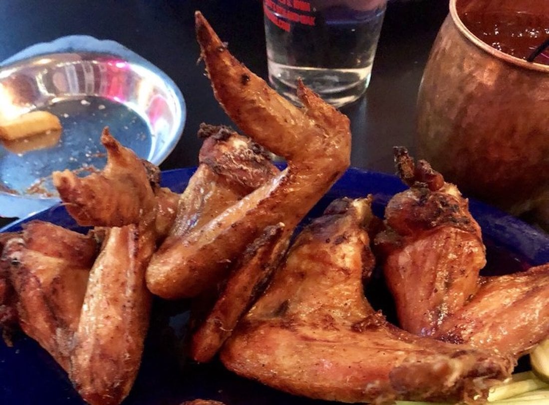 A close-up of the best chicken wings—crispy and perfectly seasoned—arranged on a blue plate, with a metal dish, a glass of water, and a copper mug in the background.