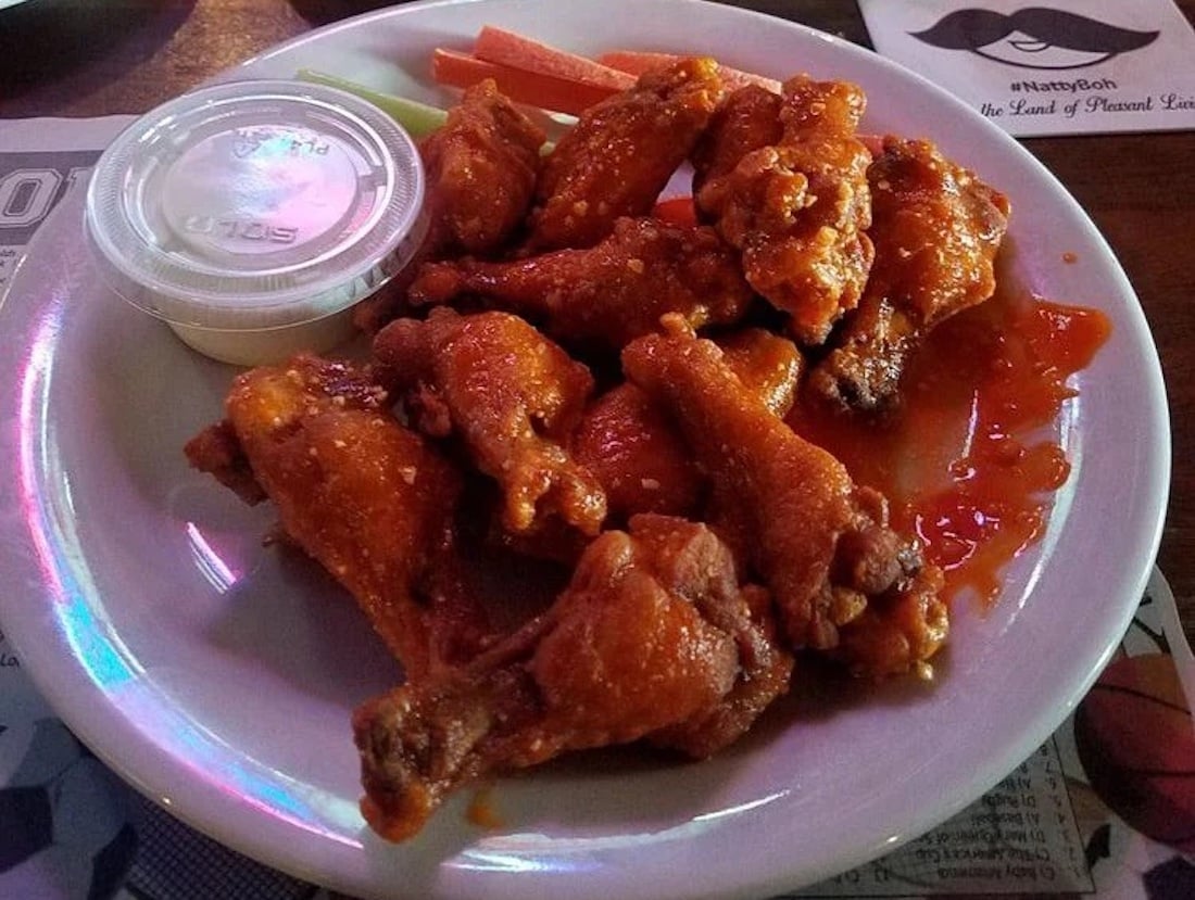 In Baltimore, Maryland, a white plate with the best chicken wings tossed in buffalo sauce, a container of dipping sauce, and a few carrot sticks on the side, all arranged on top of a placemat.