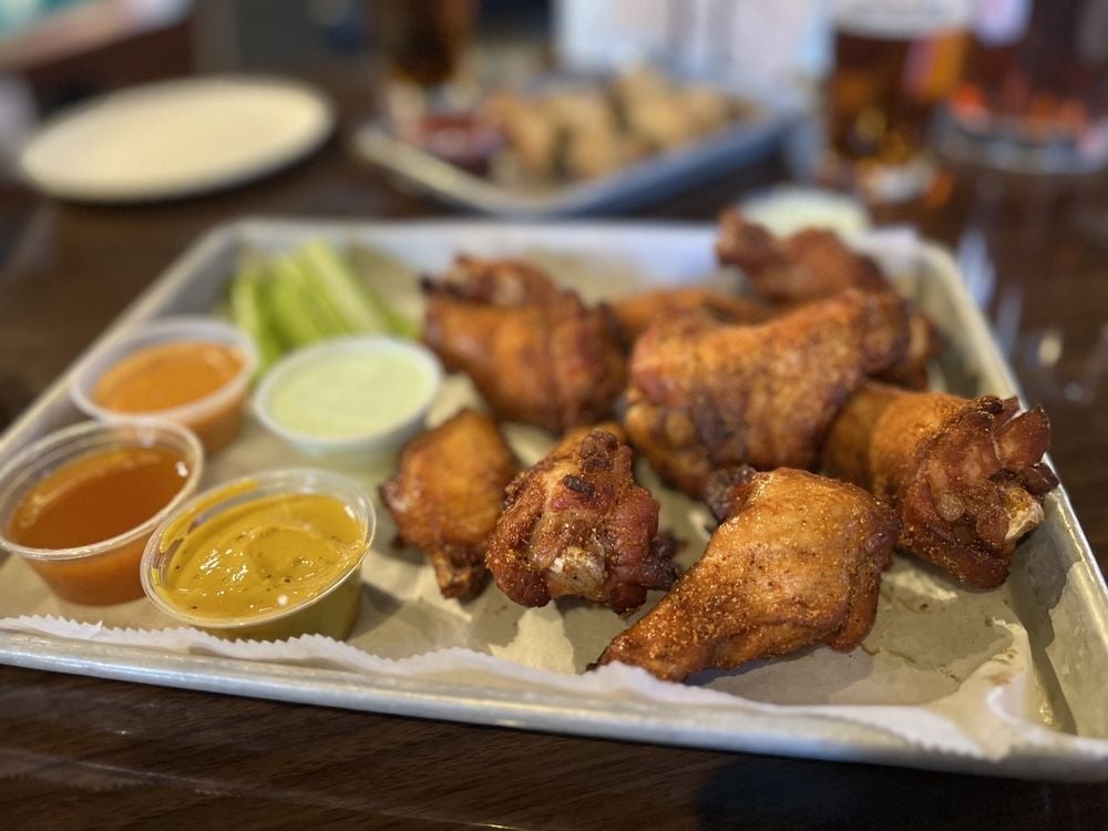A tray of the best chicken wings, perfectly crispy and served with three dipping sauces and celery sticks on the side, set on a table with blurred plates and drinks in the background.