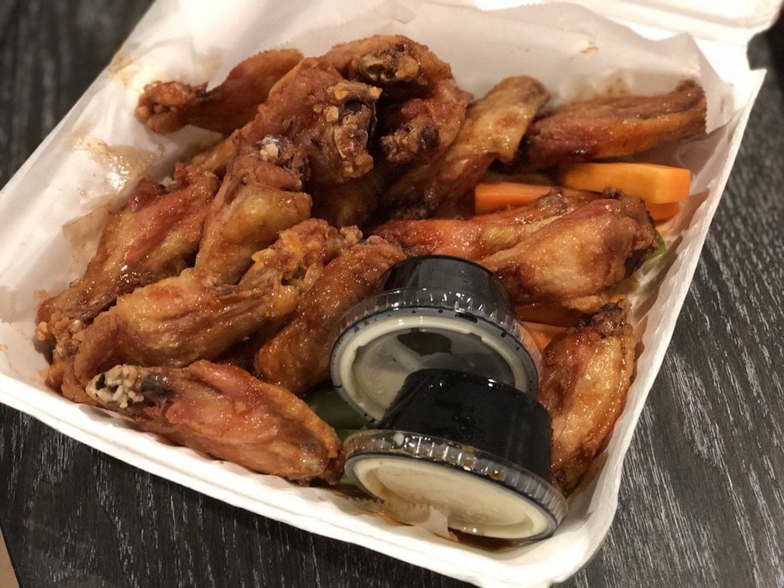 A takeout box filled with the best chicken wings—crispy and golden—served alongside carrot and celery sticks with two containers of dipping sauce. The box rests on a dark wooden surface.