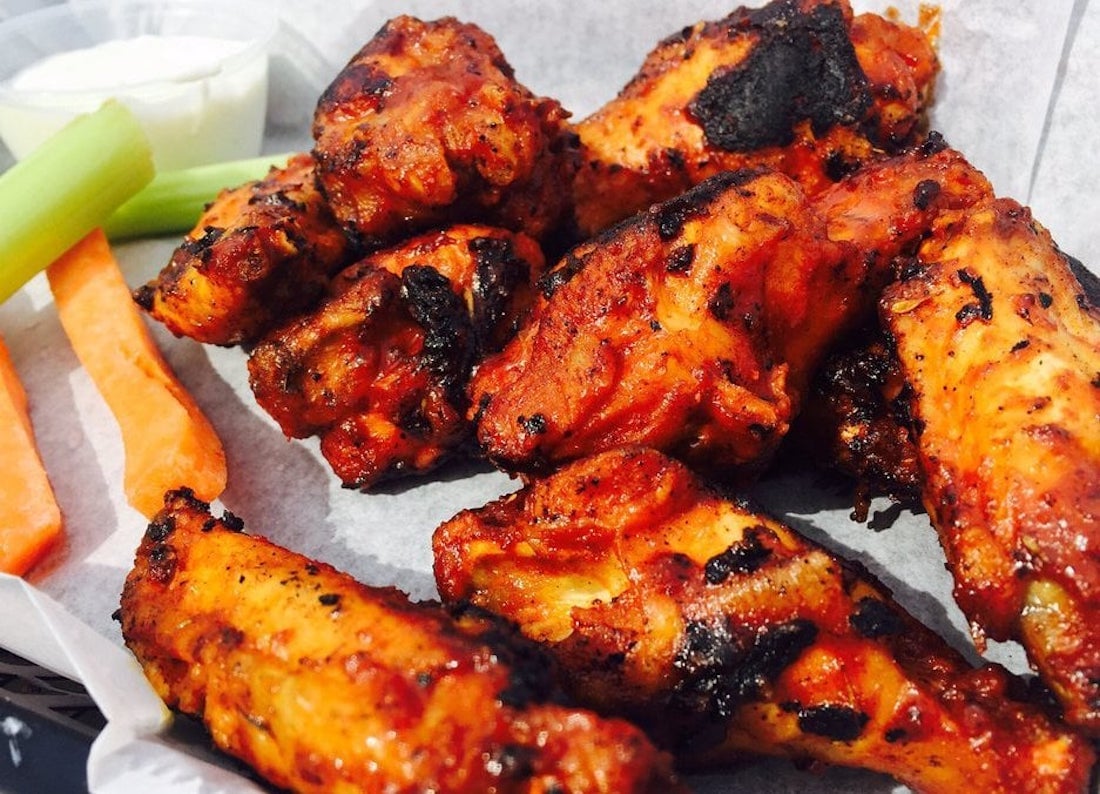 A close-up of the best chicken wings—charred and saucy buffalo style—served on parchment paper with carrot sticks, celery sticks, and a cup of ranch or blue cheese dipping sauce in the background.