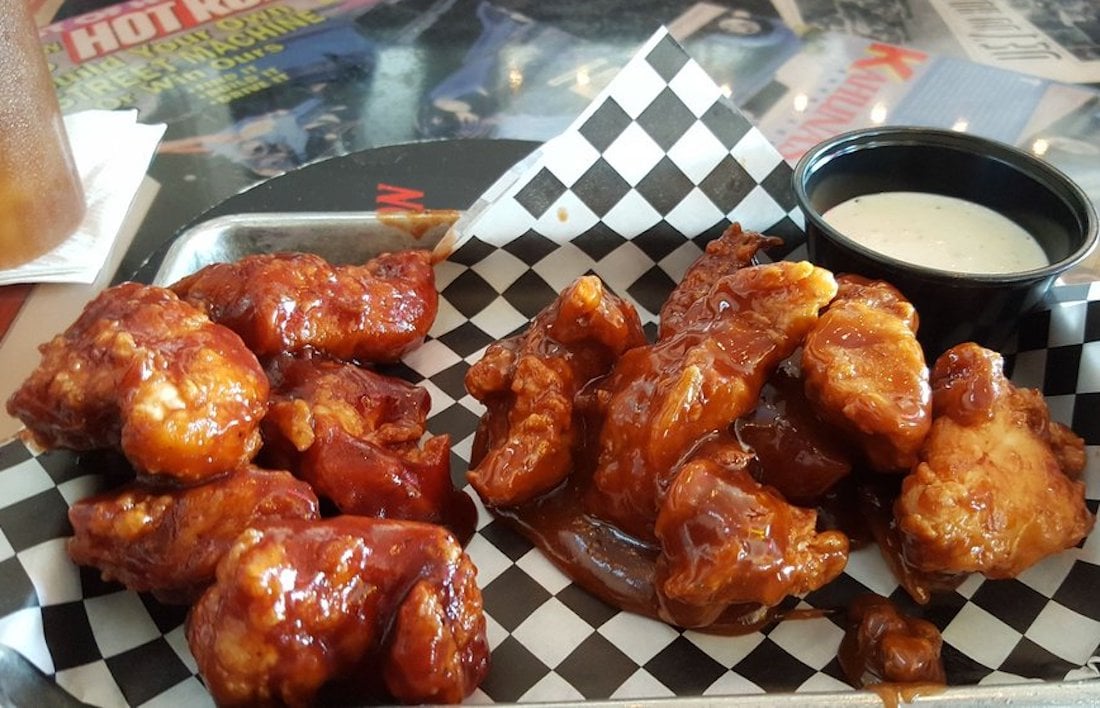 A tray of saucy, best chicken wings and boneless bites sits on checkered paper, with a small cup of ranch dressing on the side. In the background, colorful magazines add to the lively table setting.