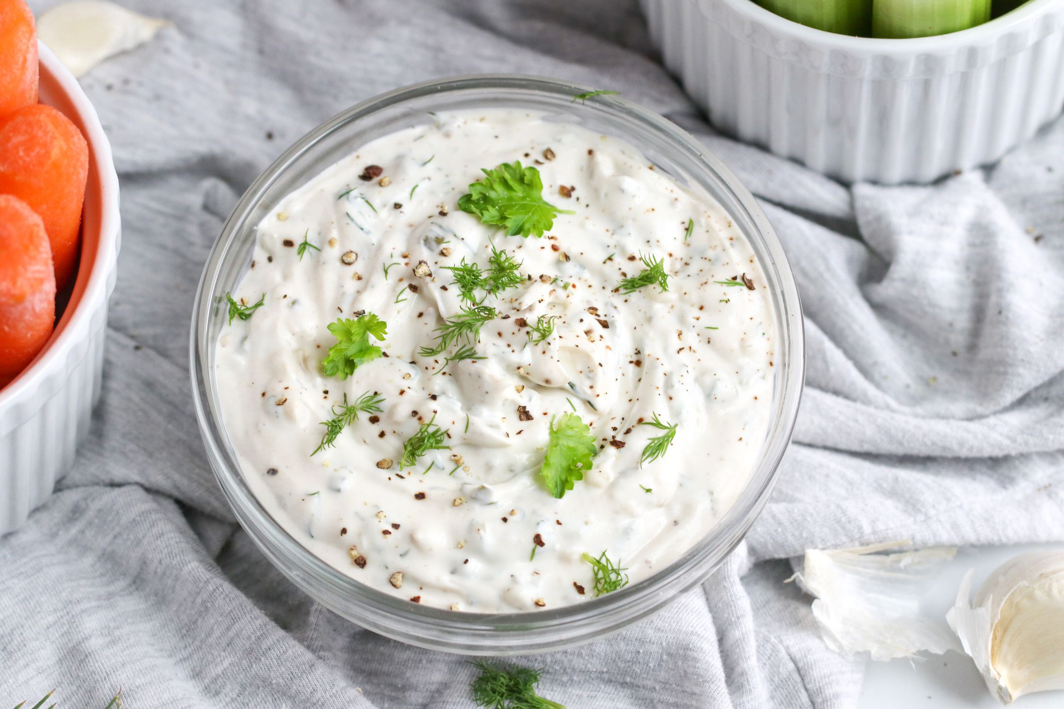 Onion dip from scratch in a glass bowl on a light grey cloth surrounded by carrots and another vegetable in white bowls