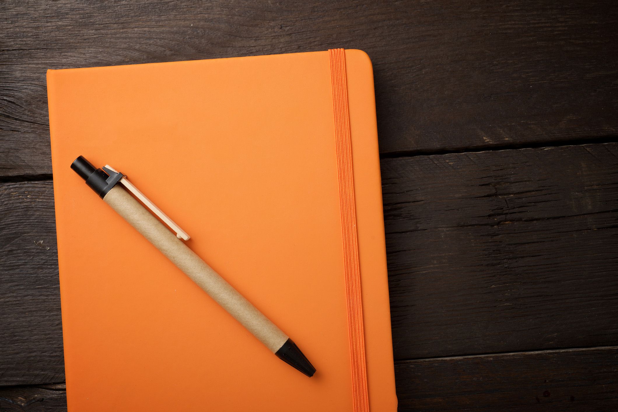 Orange closed notebook and a beige and black pen on a dark wood table