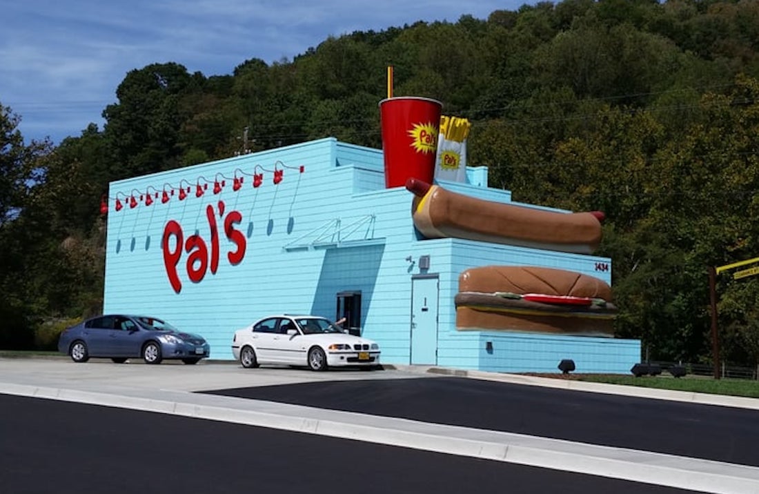 A light blue fast-food restaurant with the name "Pal's" and giant models of a hot dog, burger, fries, and drink on the roof. Two cars are parked outside, and trees are visible in the background.