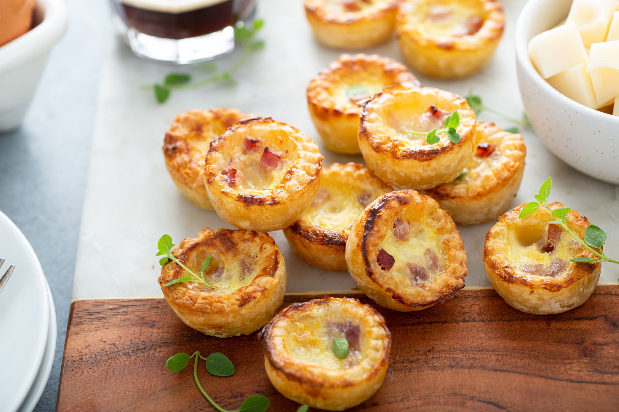 Pile of mini quiches on a wooden cutting board and a white tablecloth surrounded by plates, utensils, and other foods