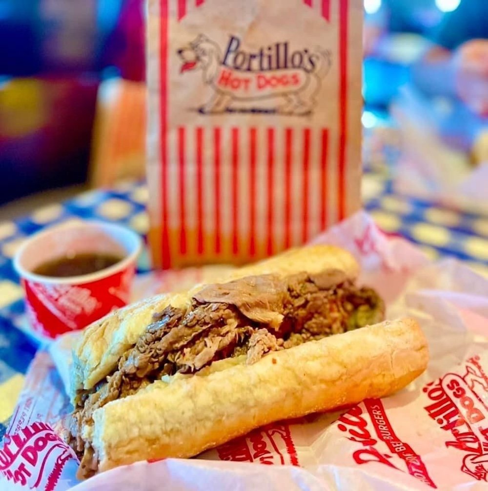 A Portillo’s Italian beef sandwich sits on branded paper, filled with juicy, sliced beef. In the background are a red-striped Portillo’s bag and a cup of au jus on a blue and white checkered table.