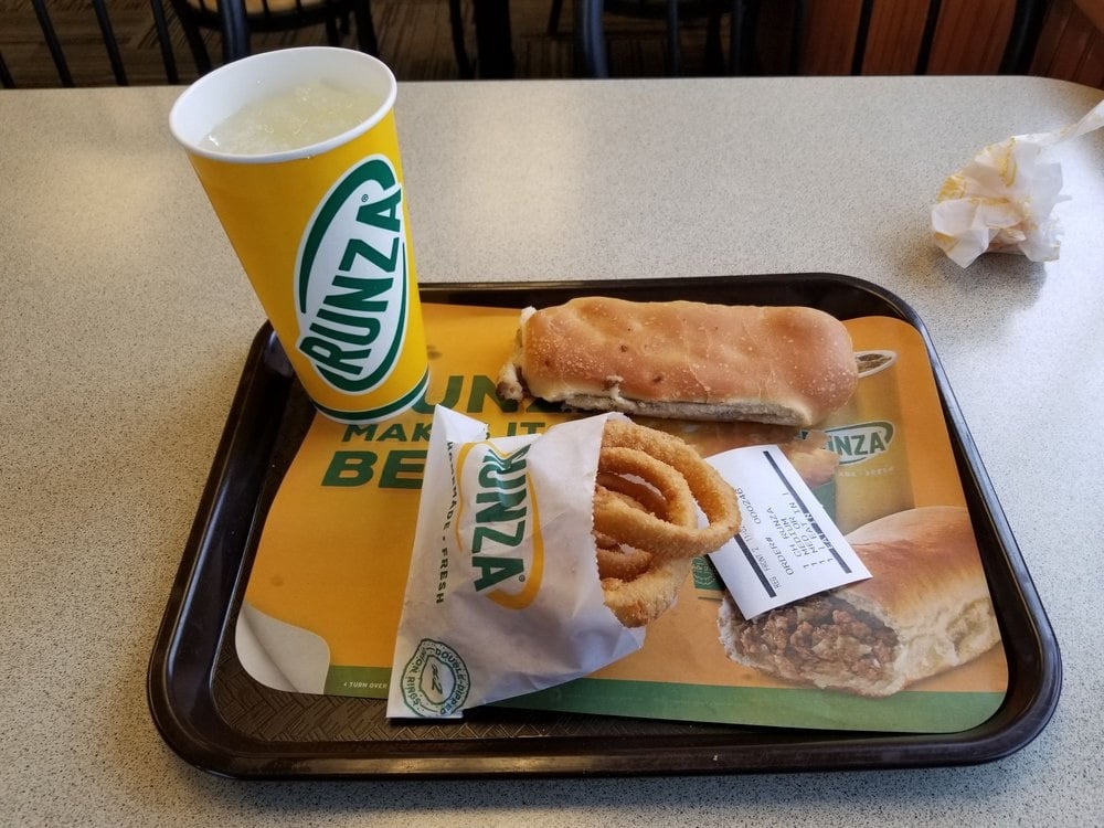 A tray with a Runza sandwich, a serving of onion rings in branded packaging, a cup of soft drink with ice, and a receipt, all on a table at a Runza restaurant.