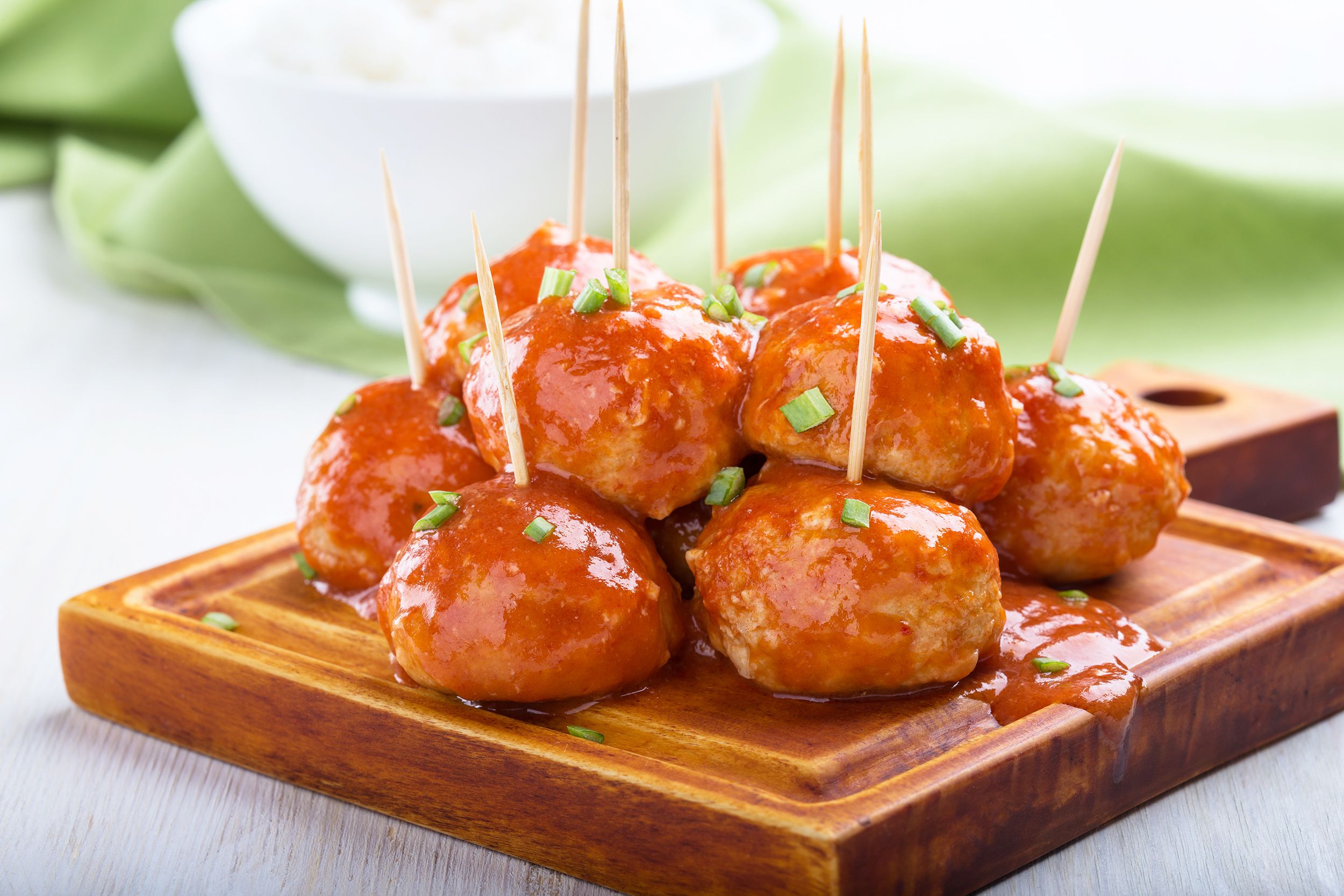 Slow cooker firecracker chicken meatballs with toothpicks on a wooden cutting board with a blurred background of a white bowl and an olive green cloth