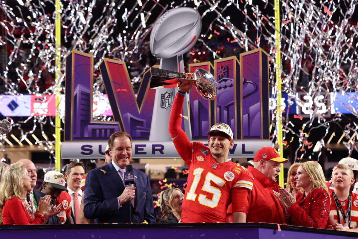 A football player in a red jersey with number 15 holds up the Super Bowl trophy on a confetti-filled stage, surrounded by teammates and media, as fans who paid high Super Bowl ticket prices celebrate under the Super Bowl LVII logo in the background.