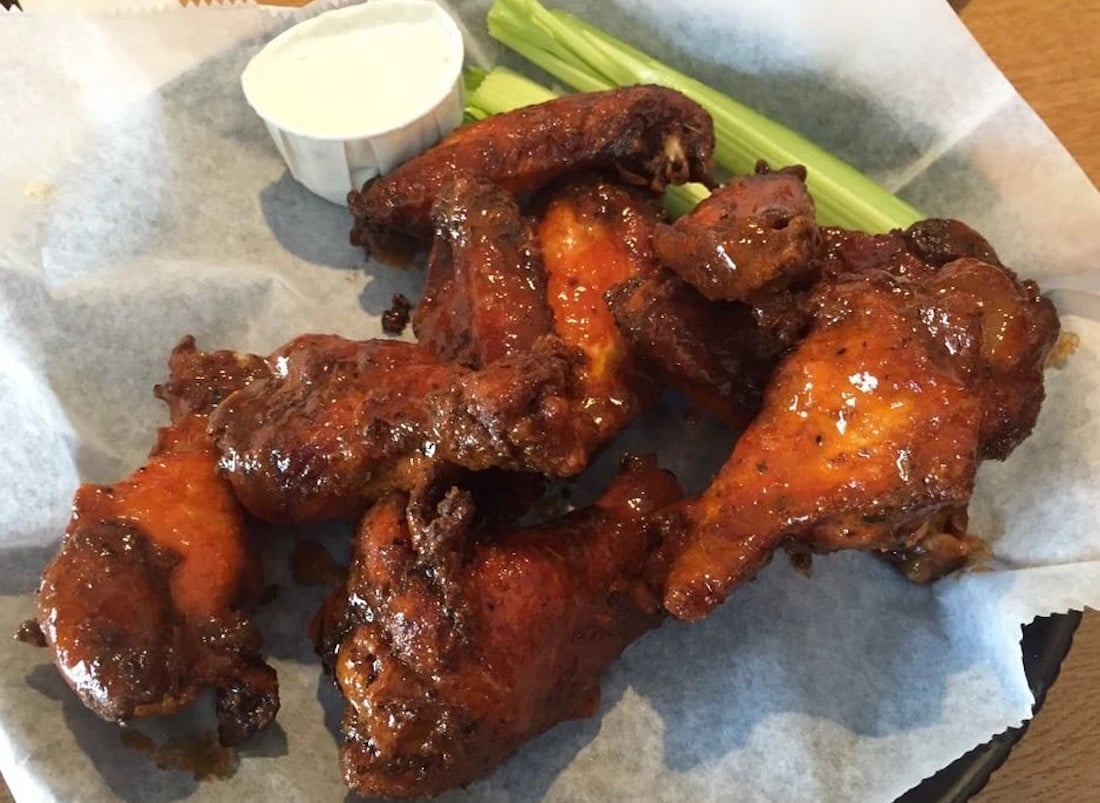 A basket of the best chicken wings, saucy and flavorful, is served with celery sticks and a small cup of ranch or blue cheese dressing on a piece of parchment paper.