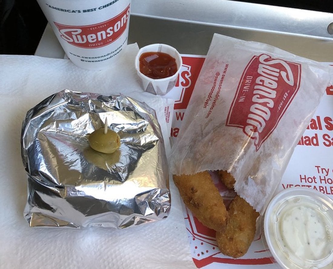 A meal from Swensons Drive-In featuring a foil-wrapped burger with a green olive on top, a bag of fried items, a cup of ketchup, a cup of ranch dip, and a drink on a tray.