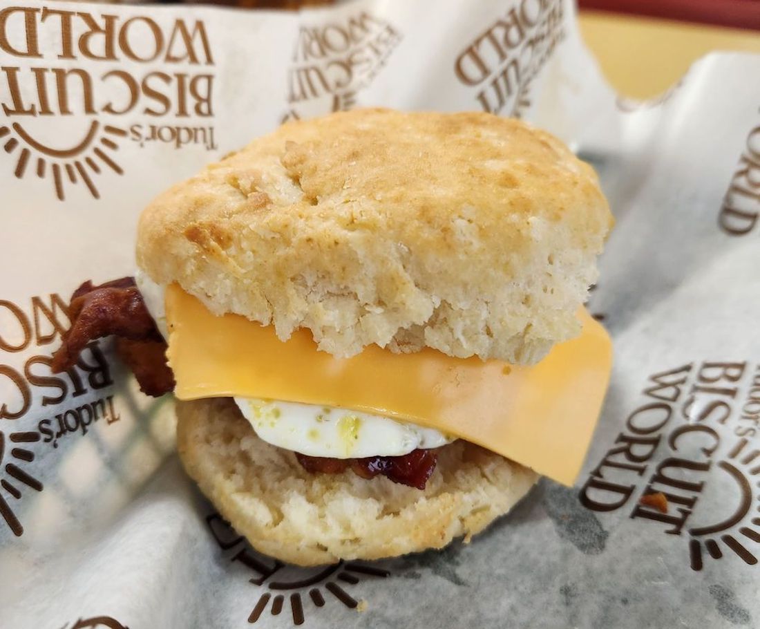 A breakfast sandwich with bacon, egg, and a slice of cheddar cheese inside a biscuit, served on branded Biscuit World paper.