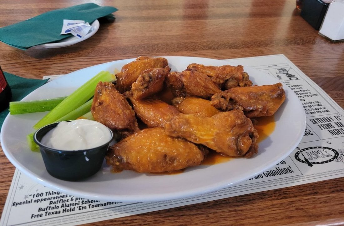 A white plate with the best chicken wings, buffalo-style, served alongside celery sticks and a cup of ranch dipping sauce on a wooden table with a restaurant menu underneath.