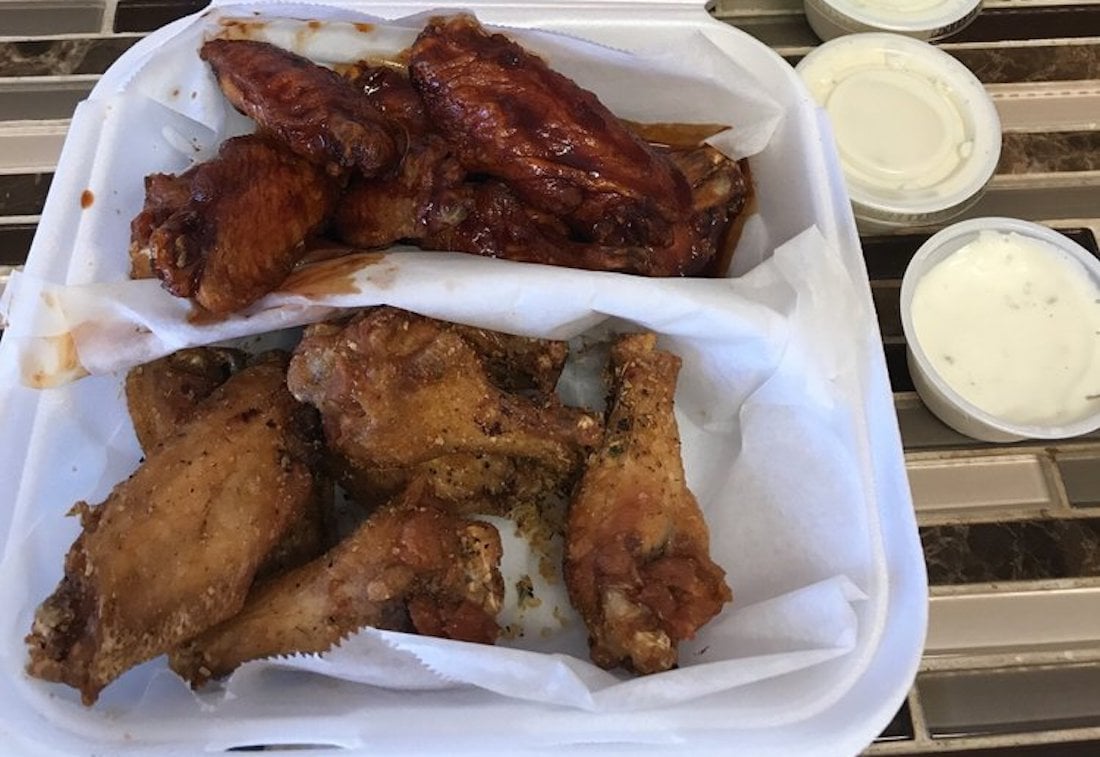 A takeout container with the best chicken wings—sauced wings on top and dry-rubbed wings on the bottom, separated by parchment paper. Three cups of dipping sauce are served on the side.