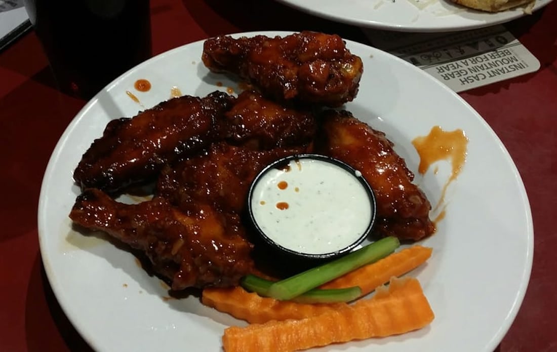 A white plate with the best chicken wings tossed in saucy buffalo sauce, a cup of ranch dipping sauce, crinkle-cut carrot sticks, and celery sticks, set on a red table.