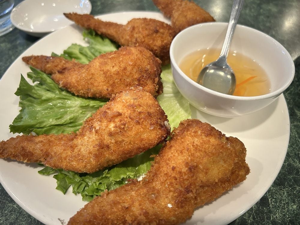 Five pieces of golden-brown fried chicken wings—truly the best chicken wings—are arranged on lettuce on a white plate, served with a small bowl of clear dipping sauce and a spoon.