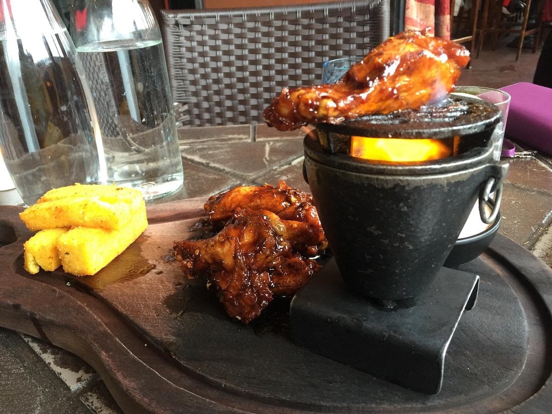 A wooden platter showcases the best chicken wings, two pieces of fried polenta, and a mini charcoal grill holding a chicken wing. Two glass water bottles and a woven chair appear in the background.