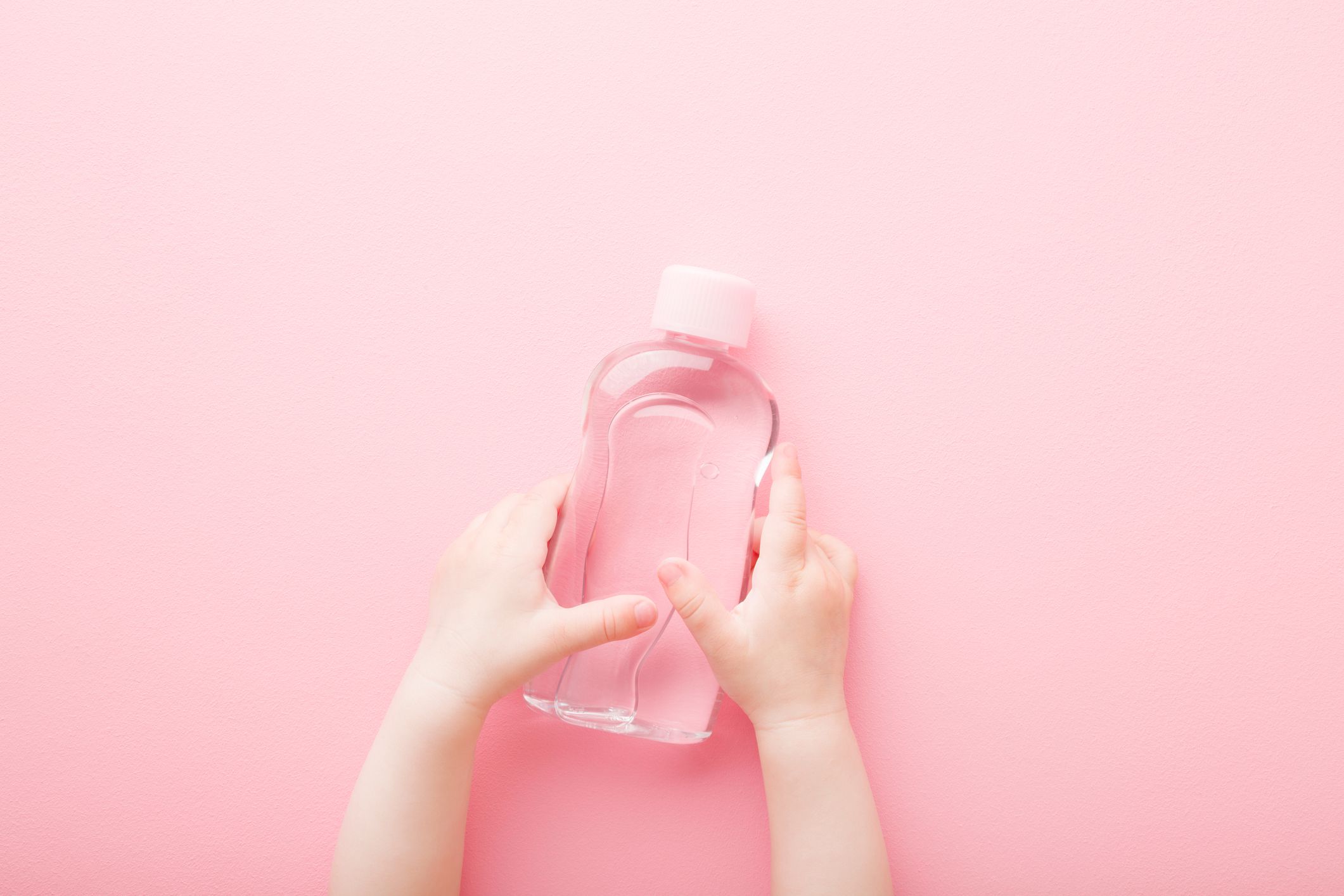 Baby girl little hands holding transparent plastic bottle of oil on light pink table background. Pastel color. Care about soft body skin. Closeup. Point of view shot. Top down view.