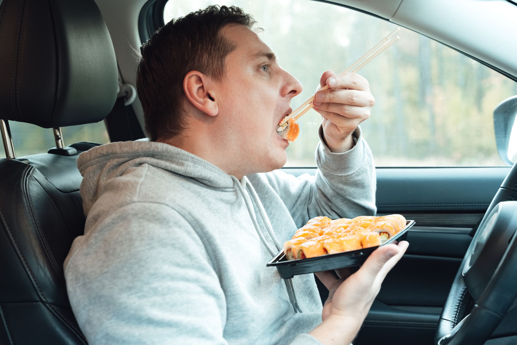 Man is eating sushi, Philadelphia rolls with fish, rice, salmon, cream cheese, avocado in car. Fast, safe, takeaway food on the road to coronavirus pandemic. Delicious japanese snacks with chopsticks.