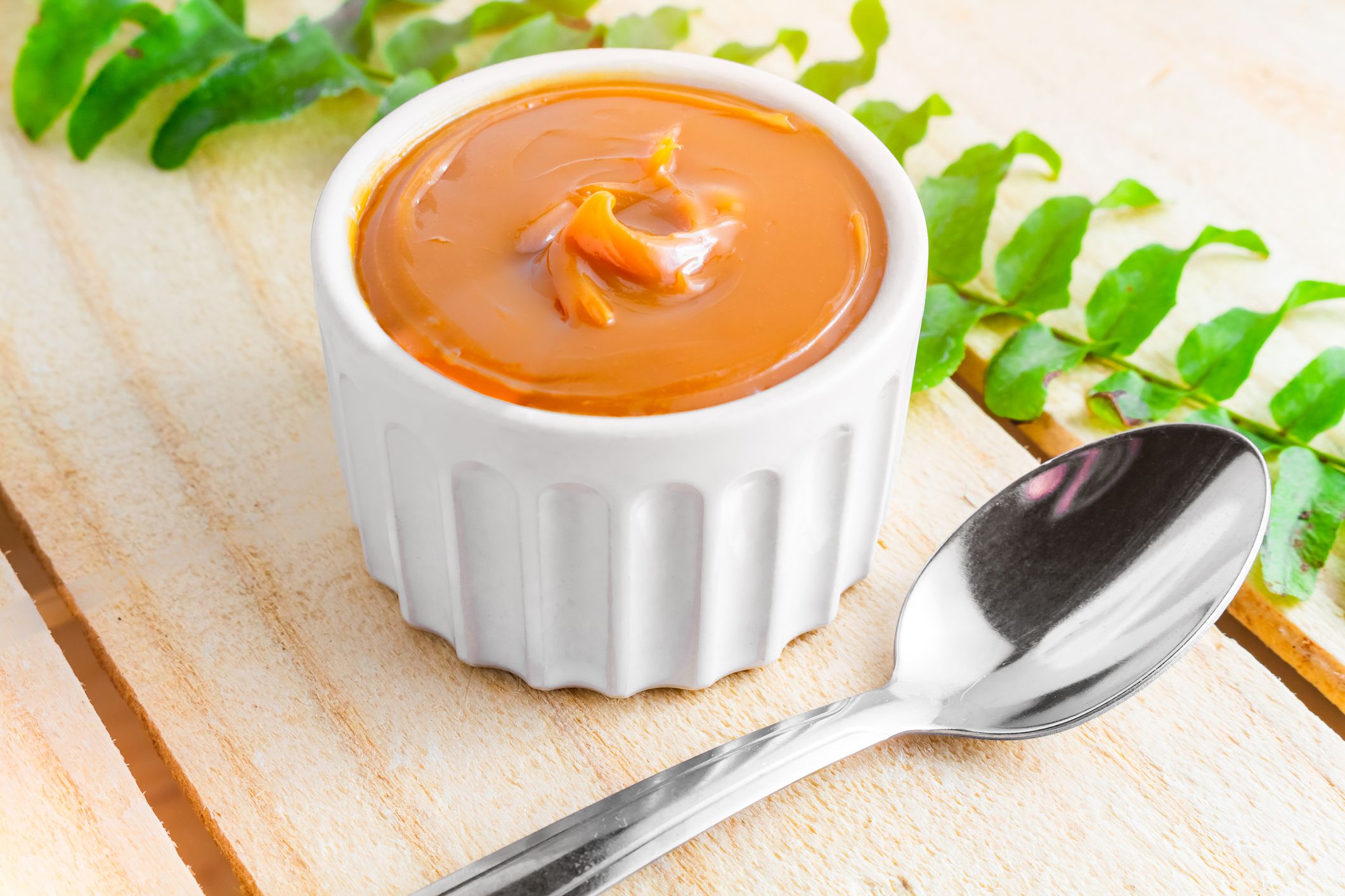 Image with a bowl of dulce de leche on the farm table, with a bunch of leaves and a spoon beside it lit by the morning sun. Typical sweet from France, Brazil and Argentina.