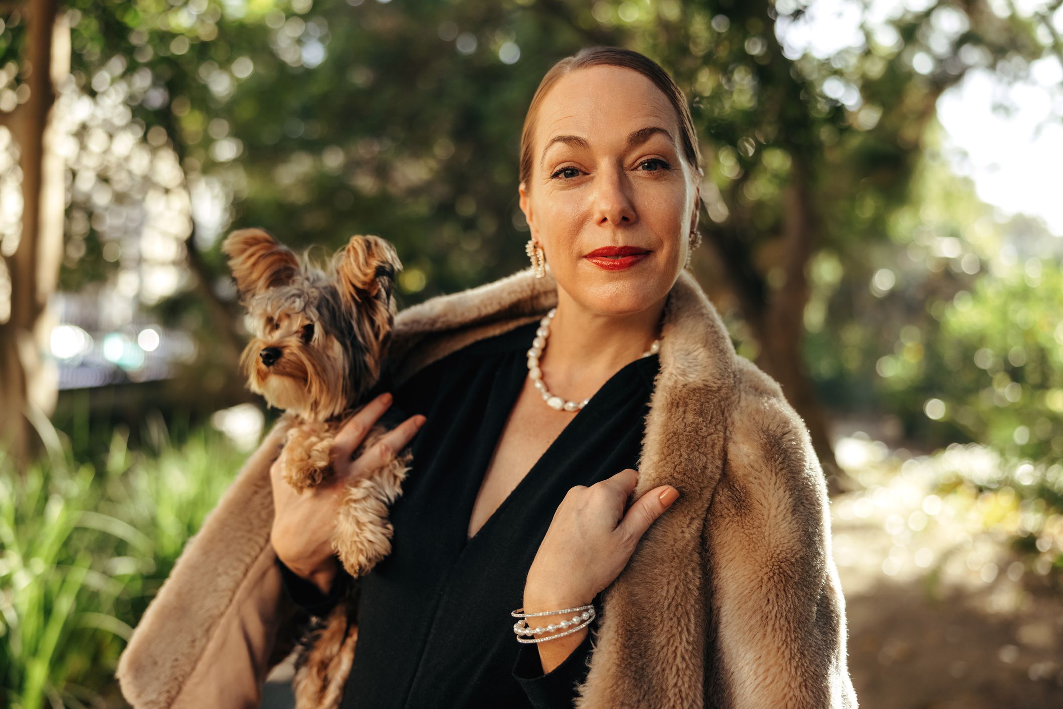 Elegant woman looking at the camera while holding her puppy outdoors. High class woman standing alone in a park during the day. Female pet owner going for a walk with her dog.