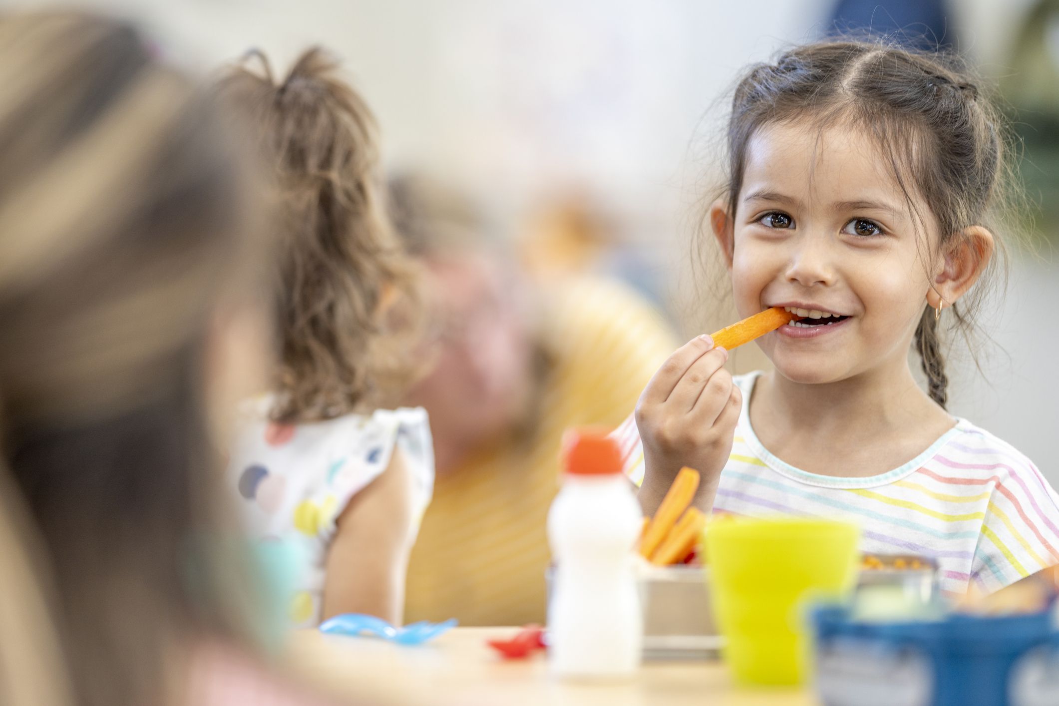 Daycare children are seen sitting around a table eating their lunch. They are dressed casually and have healthy eating choices on the table in front of them as they talk amongst themselves.