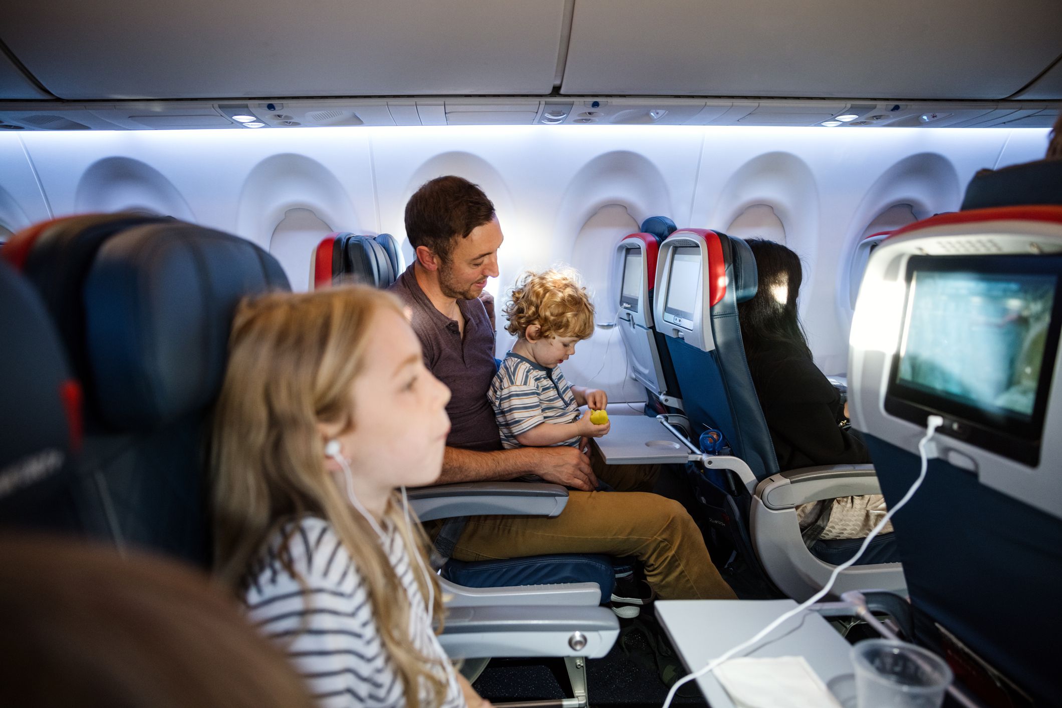 A Caucasian family with multiple children on vacation depart from the airport on a plane. The dad carries his little boy on his lap for the flight.