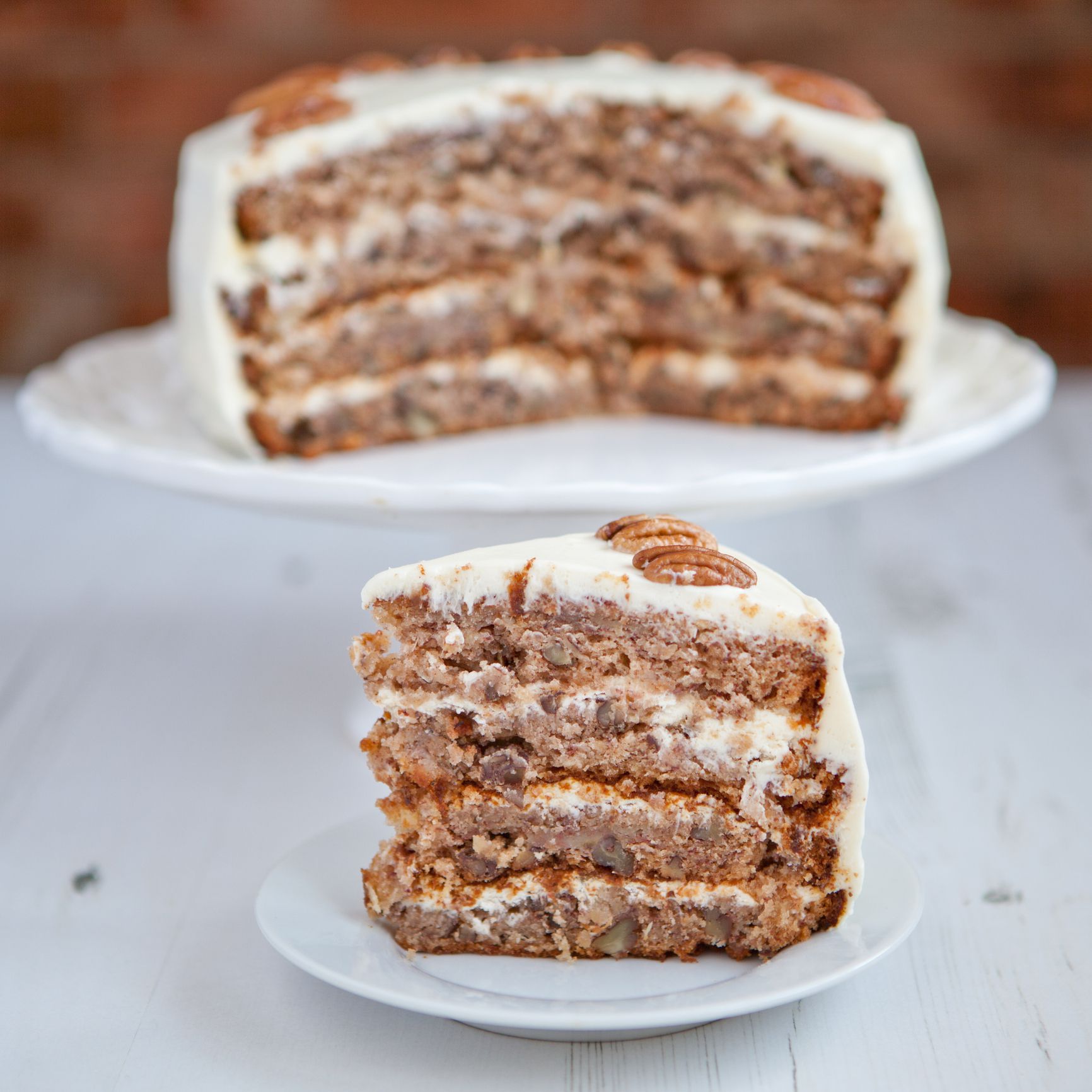 Piece of Hummingbird cake with pecans, pineapple and cream cheese frosting on the white wooden table on a brick wall background