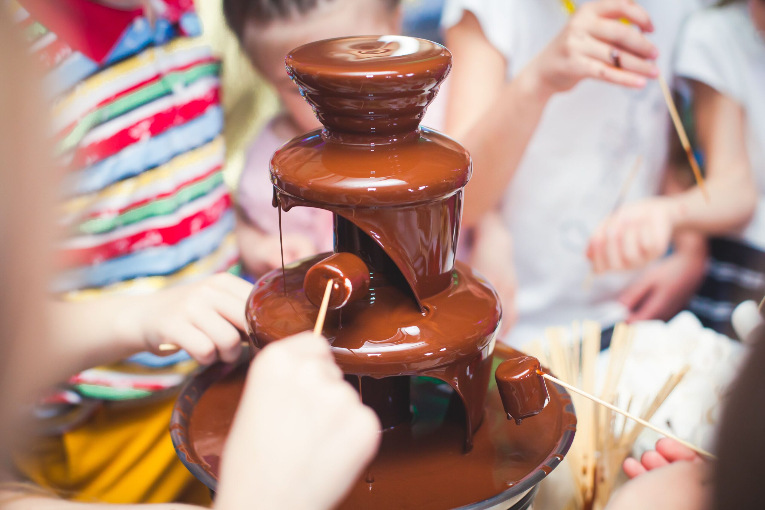 Vibrant Picture of Chocolate Fountain Fontain on childen kids birthday party with a kids playing around and marshmallows and fruits dip dipping into fountain