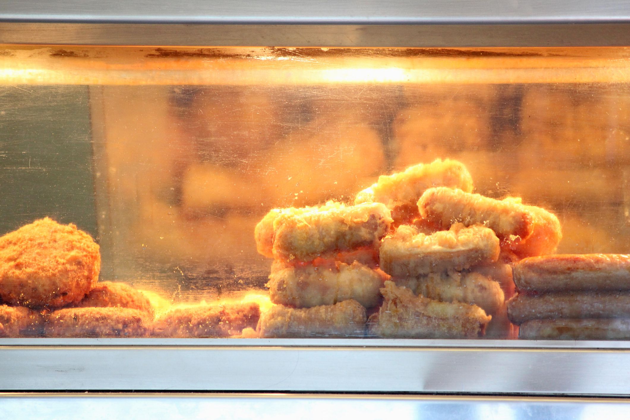 Photo showing the hot warmer cabinet in a takeaway fish and chip shop, displaying the freshly fried battered sausages, fish (cod and haddock) and fishcakes. They are keeping warm under the hot light, being visible through the glass window pane.