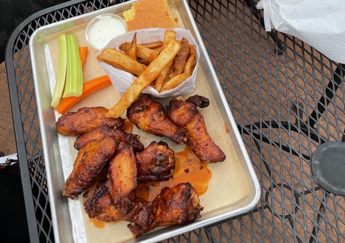 A tray with the best chicken wings in sauce, thick-cut fries in a paper cup, celery and carrot sticks, ranch dip, and a piece of cornbread, set on an outdoor metal table.