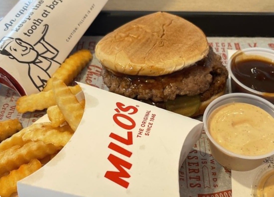 A meal with crinkle-cut fries in a Milo’s container, a burger with pickles and a soft bun, and three dipping sauces on a tray. Part of a Milo’s burger wrapper is visible in the background.