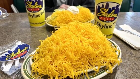 Two plates loaded with heaps of shredded cheddar cheese over Skyline Chili entrees, with Skyline cups and napkins on a table in a casual restaurant setting.