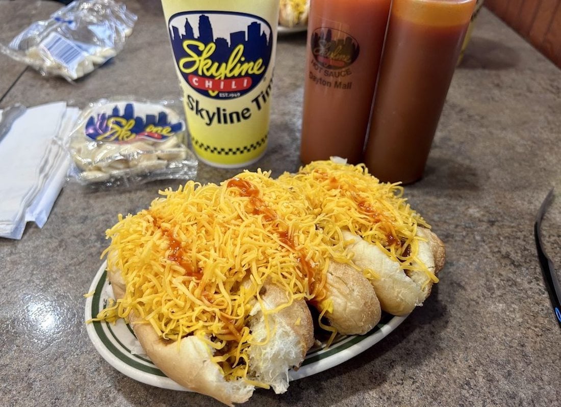 Three hot dogs topped with shredded cheddar cheese and chili are on a plate, with packets of oyster crackers, two bottles of hot sauce, and a Skyline Chili drink cup visible on a restaurant table.