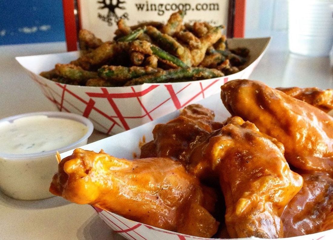 A paper tray filled with the best chicken wings, saucy buffalo style, sits next to a cup of ranch dressing and another tray of fried green beans. A Wing Coop sign is visible in the background.