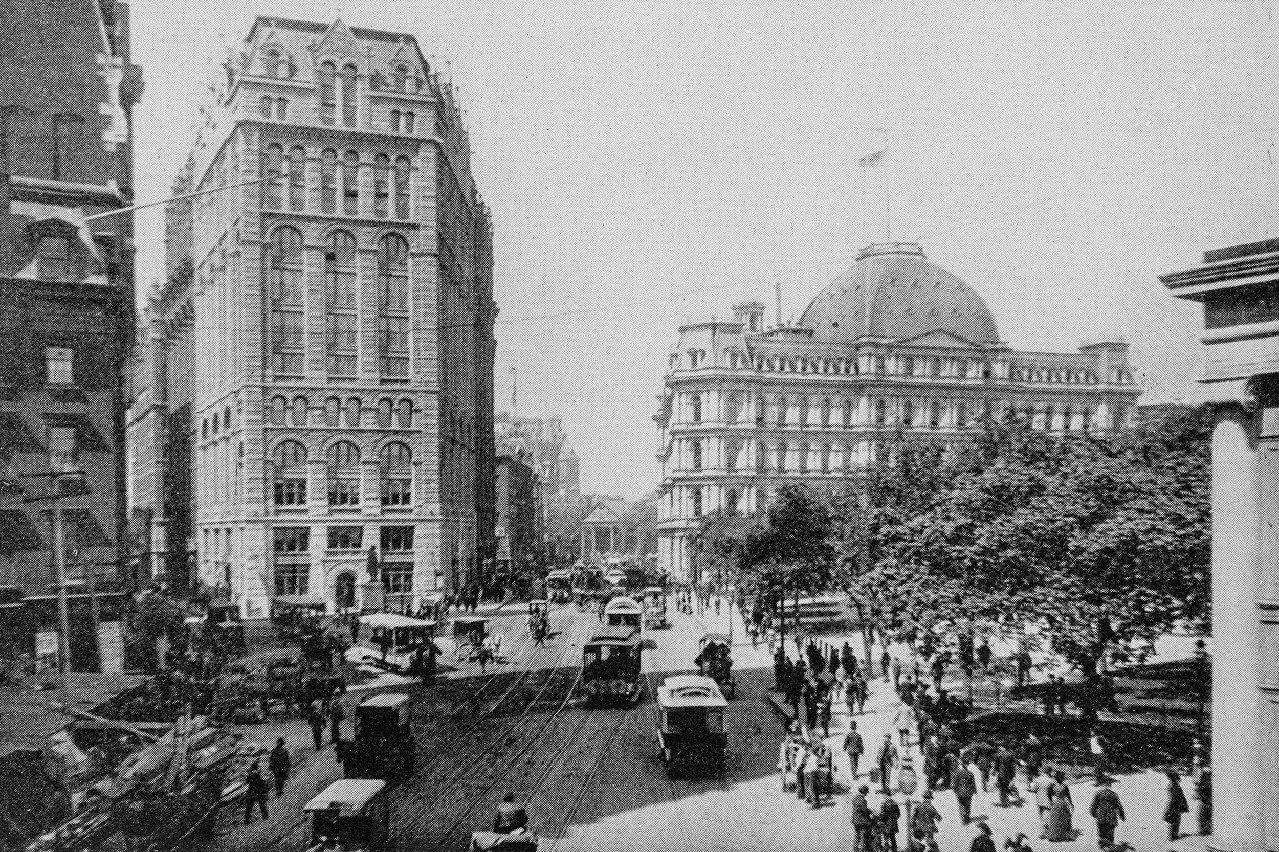 Antique black and white photograph of downtown New York City around 1910s, a park to the right and buildings on the left and in background, cable cars and people in the streets