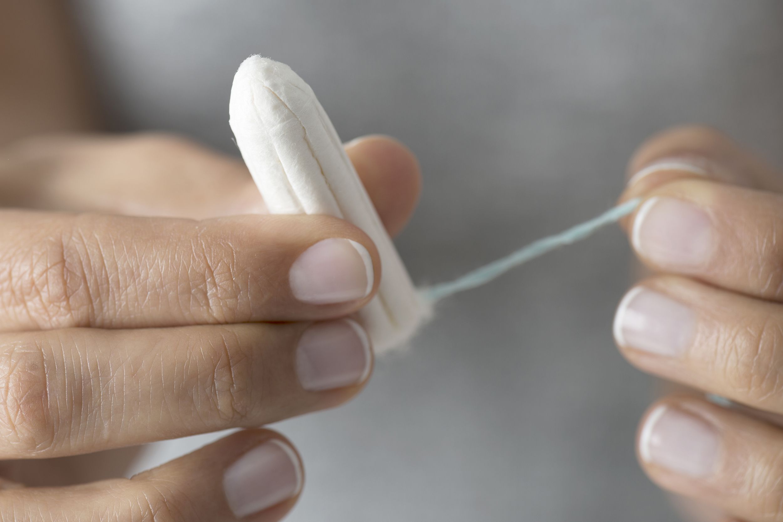 Closeup of two female hands holding tampon with blurred grey shirt background, one hand holding tampon, other hand holding string of tampon, manicured nails