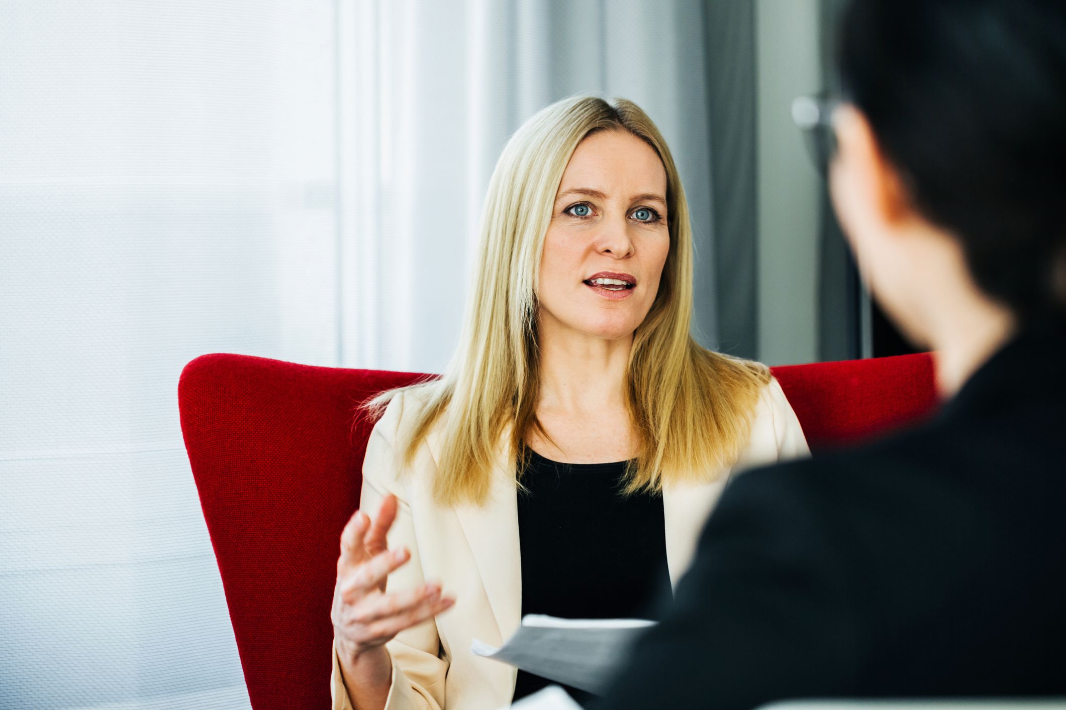 Concerned woman sitting in a red chair, discussing with coworker, selective focus, coworker blurred in the right foreground, white curtains and windows in the background