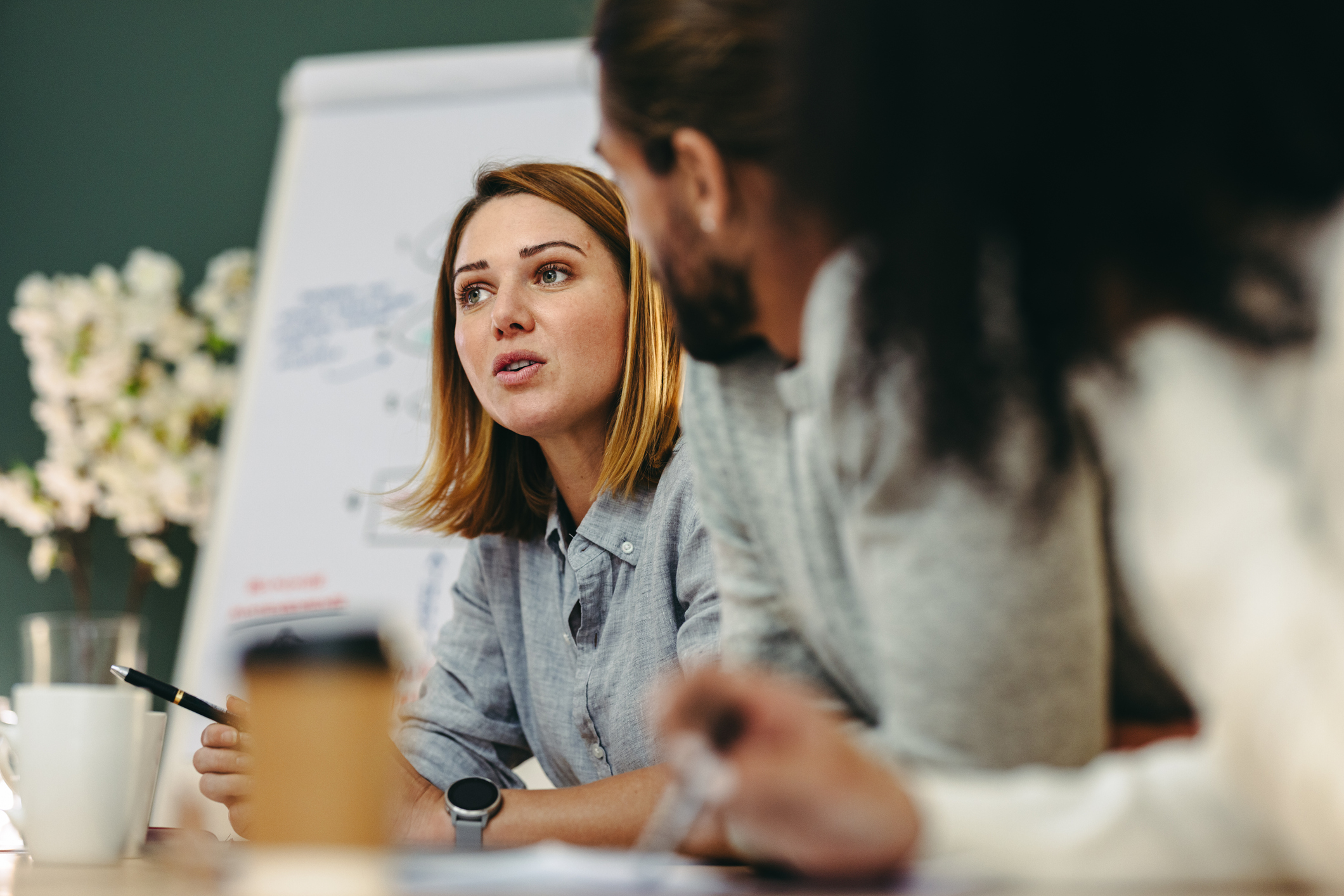 Female coworker talking at a conference table during a meeting, selective focus, two coworkers on the right blurred, white paper board and dark green wall blurred in the background