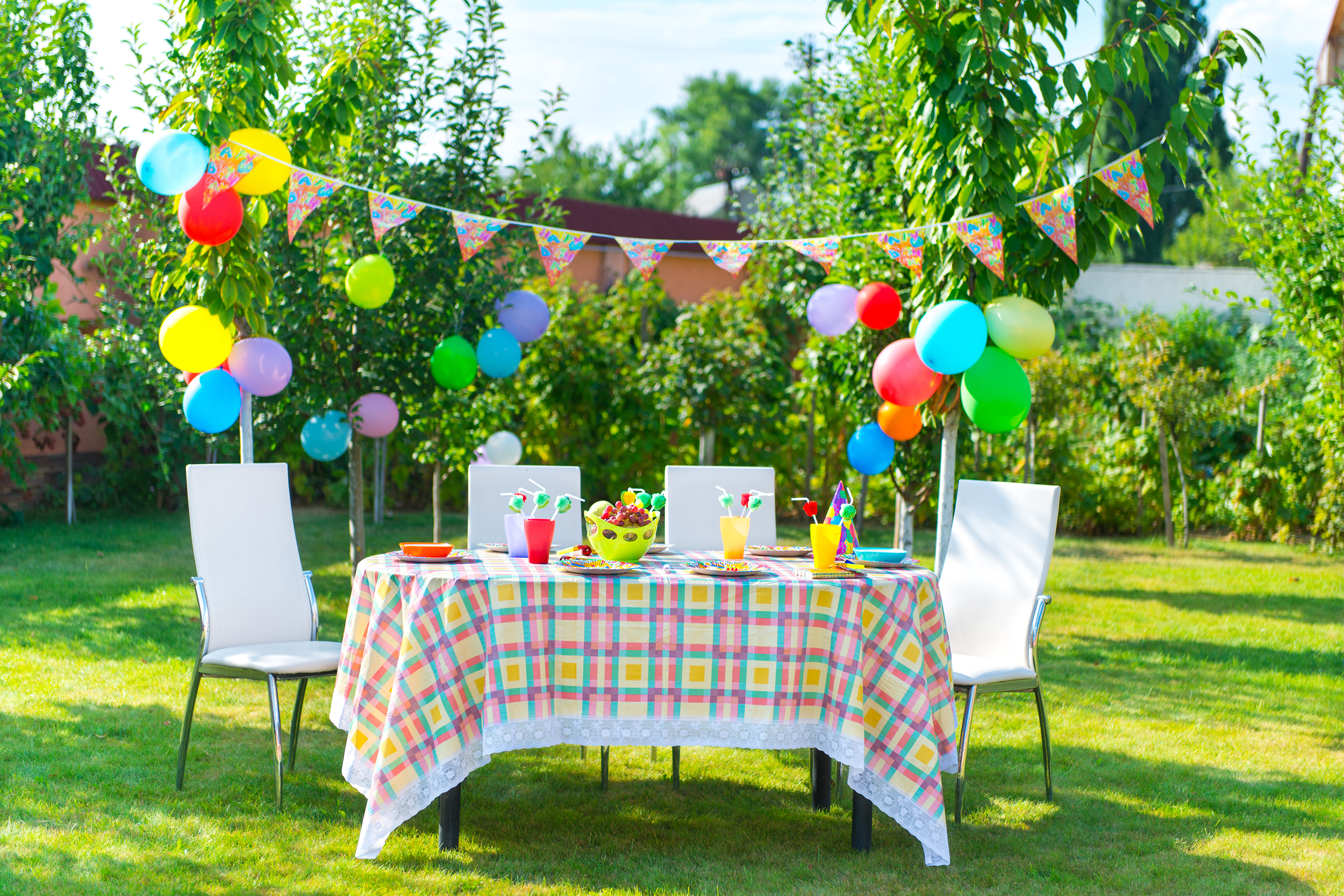 Foldable table with four white chairs, decorated for a birthday party, colorful balloons and tableware, in a yard outside, many green tree in the background, on a sunny day in summer
