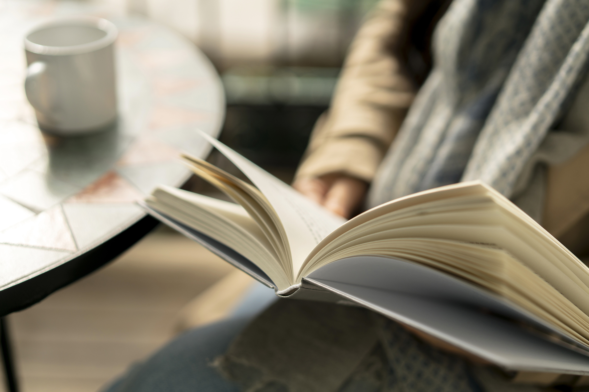Hardback book opened, being read, selective focus, in a lap of a person wearing winter clothes, sitting near a table with a white coffee mug, with a blurred background