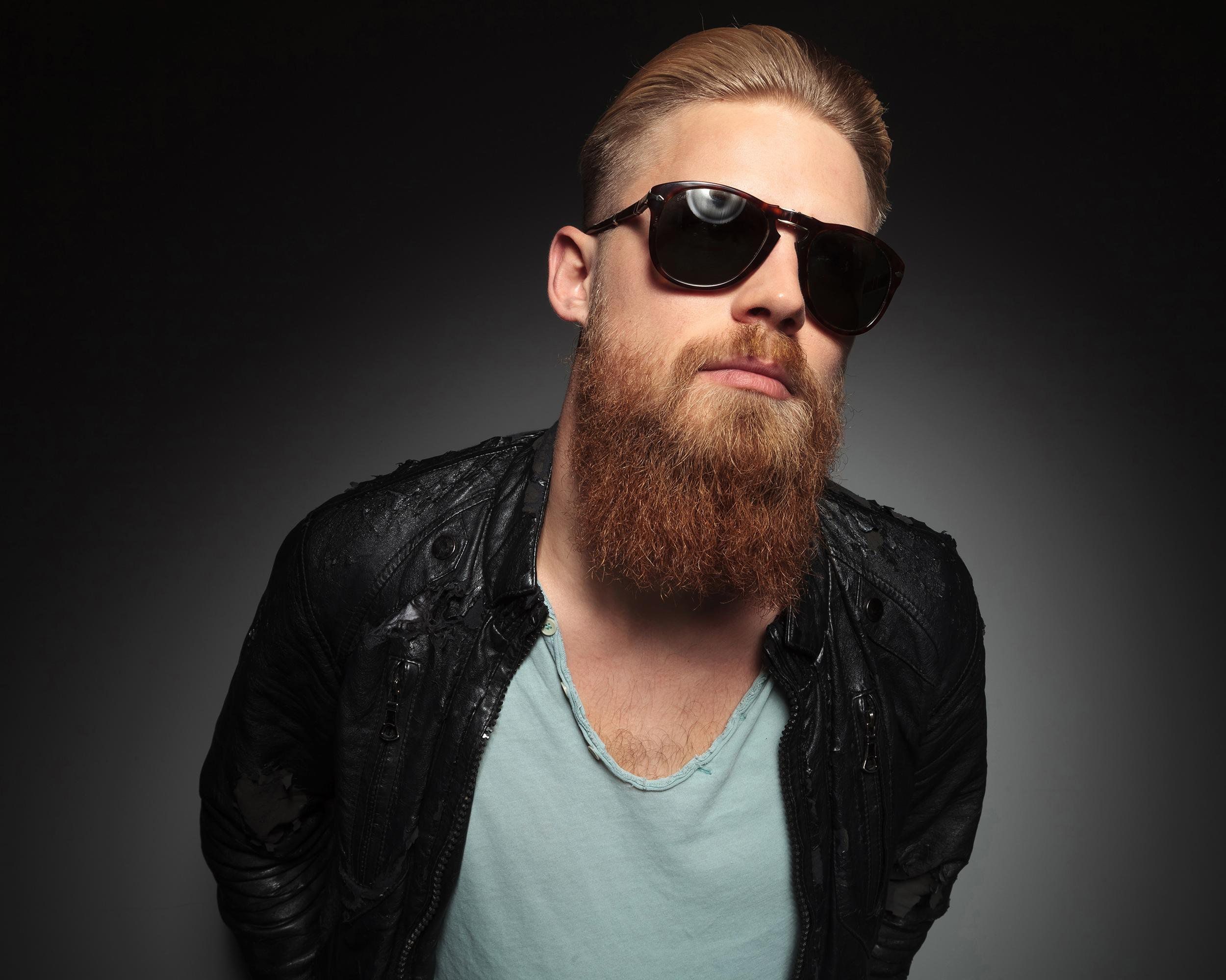 Hipster man with facial hair and beard, wearing black sunglasses, black leather jacket and grey shirt, with black gradient background