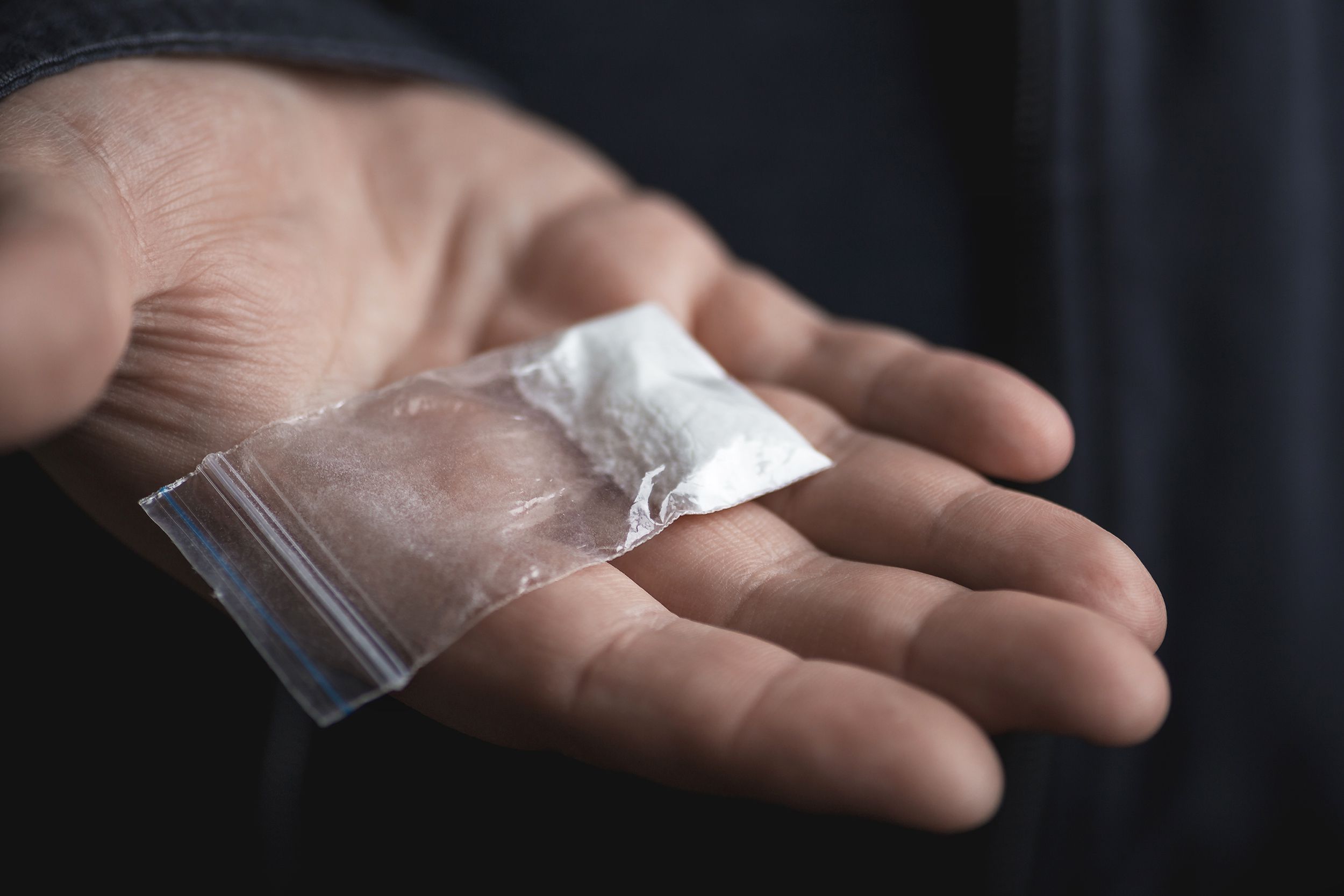 Shadowy male hand holding small bag of illegal drugs against black background